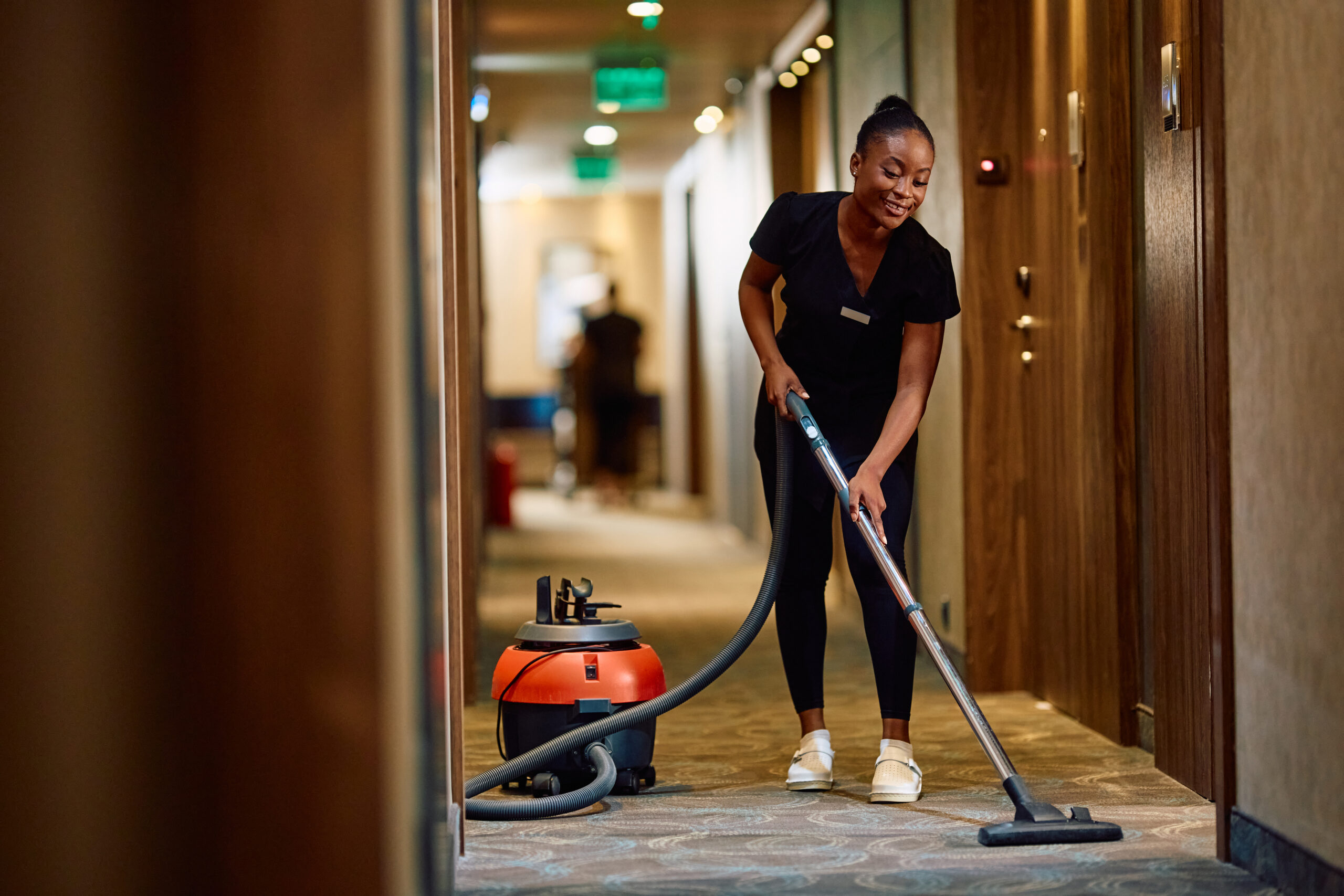 home young black maid vacuuming hallway while working in a hotel.